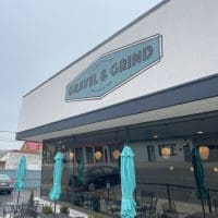 A modern cafe exterior, part of the vibrant restaurants in Frederick MD, features a sign reading Gravel & Grind above large windows. Closed teal umbrellas shade outdoor tables, with reflections of nearby buildings visible on a cloudy day.
