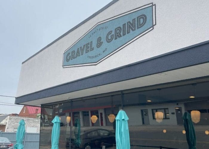 A modern cafe exterior, part of the vibrant restaurants in Frederick MD, features a sign reading Gravel & Grind above large windows. Closed teal umbrellas shade outdoor tables, with reflections of nearby buildings visible on a cloudy day.