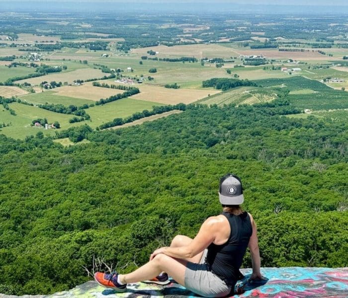 A person in a black tank top, gray shorts, and a cap sits on a colorful graffiti-covered rock—one of the best hikes around Frederick MD—taking in sweeping views of farmland, fields, distant hills, and lush green forest under a clear sky.