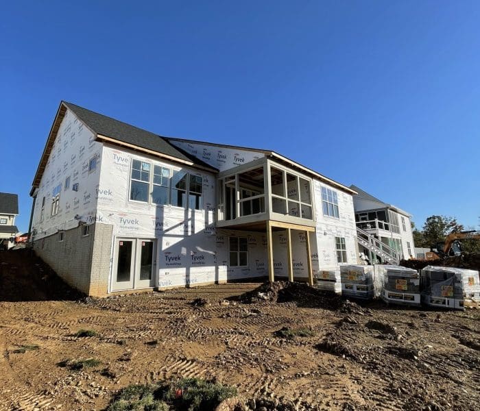 Two‑story house under construction with Tyvek wrap, exposed framing, and a dirt lot in the foreground while the sky is clear blue.