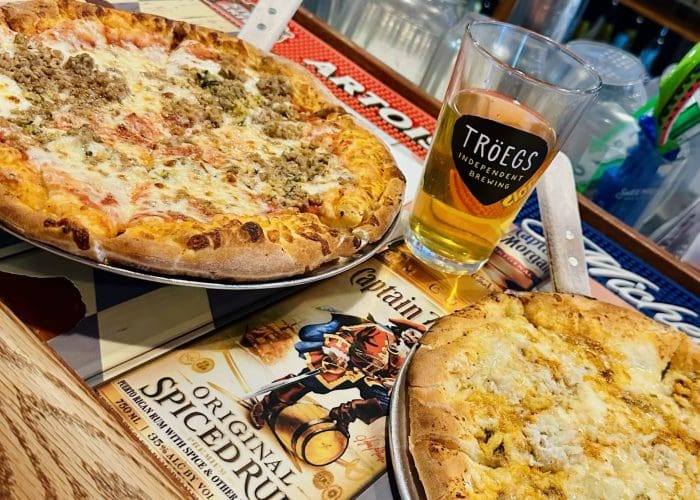 Two pizzas sit on metal trays atop a bar counter in pizza in Fredrick MD. One features sausage and cheese, the other is a garlicky white pizza. A pint of Tröegs beer sits between them, with bar mats and drink bottles visible in the background.