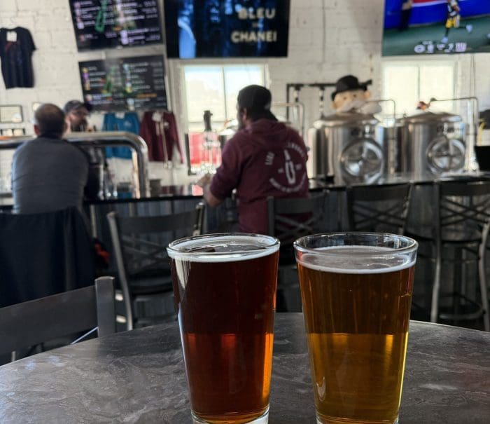 Two pint glasses of beer—one dark amber, one light golden—sit on a round table in a Mt Airy MD brewery. In the background, people relax at the bar with stainless steel brewing tanks, TV screens, and a menu board on a white brick wall.