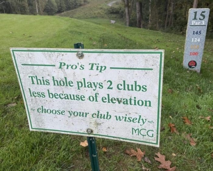 A golf course tee in Mt Airy MD features a sign: “This hole plays 2 clubs less because of elevation—choose your club wisely.” To the right, a stone marker notes Hole 15, Par 3, with distances listed; trees and fairway stretch into the background.