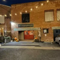 A brick Bar with string lights overhead features a sign reading McClintock's Back Bar above red double doors. Pumpkins sit by the entrance, with a barrel and umbrella nearby. It's nighttime, hinting at the secret charm inside while cars line the sides.