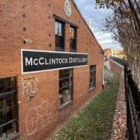 A red brick building in Frederick, MD with a black-and-white “McClintock Distillery” sign. Barred windows, a rooftop clock, graffiti, and autumn leaves by a black metal fence make it one of the unique things to do in Frederick. The sky is partly cloudy.