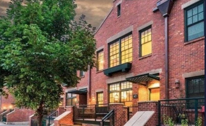 A row of modern brick townhouses at dusk, with large lit windows and black railings lining small front steps—these inviting homes evoke the charm you'd seek when deciding where to stay in Bend, Oregon. A leafy tree completes the picturesque scene.