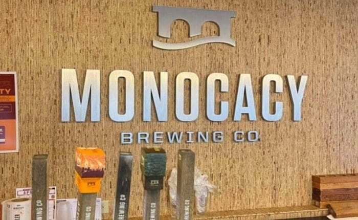A wall with the large silver letters “MONOCACY BREWING CO” and a bridge logo rises above a modern bar counter. Five taps labeled with “Monocacy Brewing Co.” hint at why it’s among the best breweries in Frederick MD. Shelves below hold clean glassware.