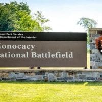 A large stone and metal sign on grass reads Monocacy National Battlefield, a top highlight among things to do in Frederick, MD. The National Park Service emblem is featured, with trees and a white building with a green roof in the background.
