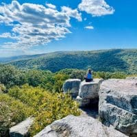 A person sits on a large rock, facing away, overlooking a vast, green forest with rolling hills under a bright blue sky. Sunlight highlights the rocky ledge and lush treetops—an inspiring scene from one of the best hikes around Frederick MD.