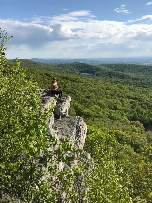 A person sits on the edge of a rocky cliff overlooking a vast green forest, one of the best hikes around Frederick MD. Trees cover rolling hills to the horizon, with a small distant lake nestled in the landscape under a partly cloudy sky.