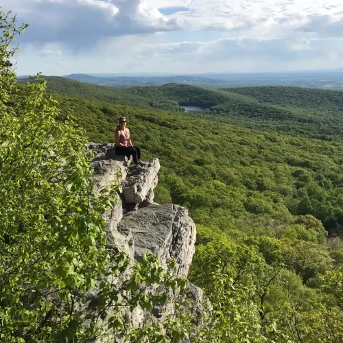 A person sits on the edge of a rocky cliff overlooking a vast green forest, one of the best hikes around Frederick MD. Trees cover rolling hills to the horizon, with a small distant lake nestled in the landscape under a partly cloudy sky.