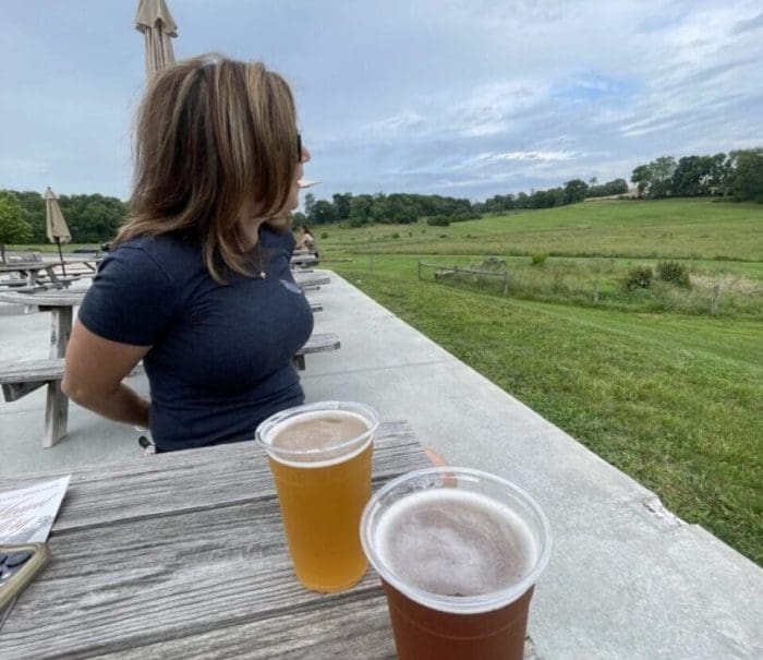 A woman with shoulder-length hair sits at a wooden picnic table outdoors in New Market, MD, gazing toward a green field under a cloudy sky. Two plastic cups of beer rest on the table, with umbrellas and more tables visible in the background.
