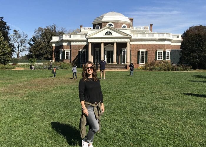 Woman in black long-sleeve top and jeans posing on a grassy lawn in front of a large brick neoclassical building with columns and a dome, under a clear blue sky.