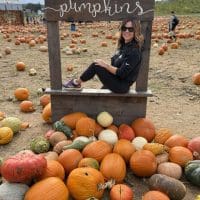 A woman with wavy brown hair, wearing sunglasses, a black outfit, and purple shoes, sits smiling in a wooden frame labeled pumpkins at Gaver Farm. She is surrounded by orange and multicolored pumpkins at a lively pumpkin patch.