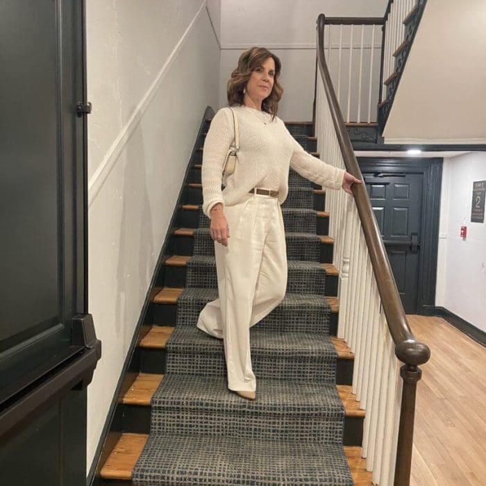 A woman with wavy brown hair, wearing a cream sweater and wide-leg white pants, stands midway on a carpeted staircase indoors—her style echoing the modern, clean interior design found in boutique hotels Frederick MD is known for.