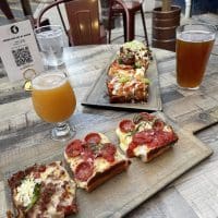 Two wooden boards hold Detroit-style pizza slices with toppings like pepperoni, sausage, jalapeño, and vegetables. Nearby, two beers and a QR code menu add to the vibe of enjoying pizza in Fredrick MD at this inviting outdoor dining spot.