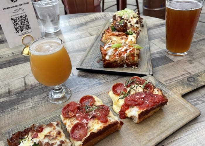 Two wooden boards hold Detroit-style pizza slices with toppings like pepperoni, sausage, jalapeño, and vegetables. Nearby, two beers and a QR code menu add to the vibe of enjoying pizza in Fredrick MD at this inviting outdoor dining spot.