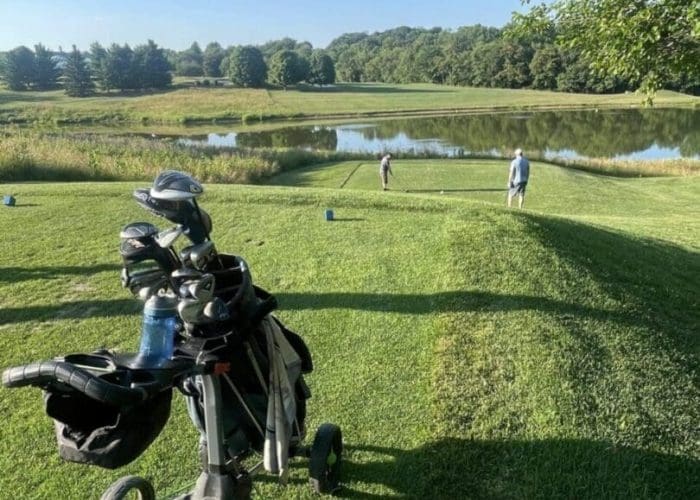 A golf bag on a push cart sits on green grass in the foreground. Two golfers near a tee by a pond, surrounded by trees and under a clear blue sky, enjoy a sunny day at the Mt Airy MD course.