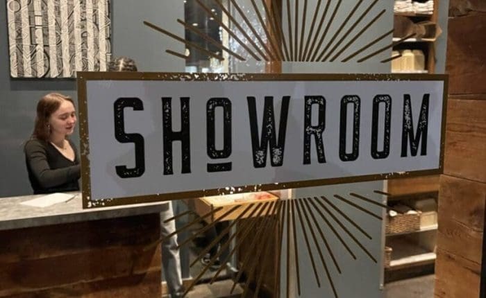 A glass door with a large “SHOWROOM” sign and gold rays welcomes you inside. A woman stands behind a wood reception desk, shelves display items, and the dark gray walls echo the stylish decor found in restaurants in Frederick MD.