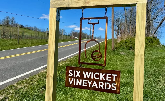 A wooden frame holds a metal sign for “Six Wicket Vineyards” by a rural roadside. Known among Frederick MD Wineries, the sign features a stylized emblem above the name, set on a grassy slope with fences, trees, and blue sky in the background.