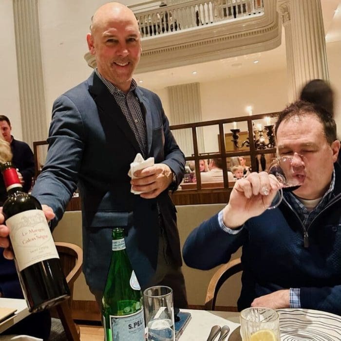 A smiling server in a suit presents a bottle of red wine to a guest at the best steakhouse Frederick MD, where tall white columns frame the restaurant. The table features San Pellegrino, a cocktail, and a plate with a napkin.