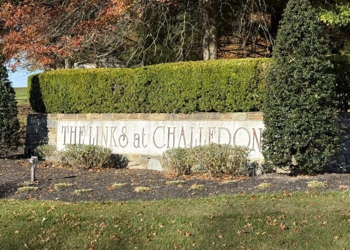 A stone sign with neatly trimmed hedges and two tall bushes displays the words The Links at Challedon. Autumn leaves and sunlight add warmth to the scene, while a small light fixture highlights the Links at Challedon's welcoming entrance.