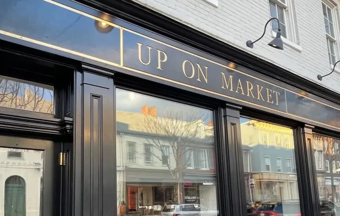 A storefront with black trim and large windows reflects nearby buildings and cars. Above, a gold-trimmed black sign reads UP ON MARKET, one of the best bars in Frederick. The white brick facade features potted plants outside.