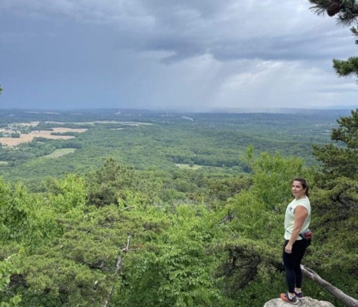 A woman in athletic clothes stands on a rocky ledge overlooking a vast green forest, one of the best hikes around Frederick MD. She gazes at the camera, framed by dramatic scenery and sunlight breaking through distant clouds.
