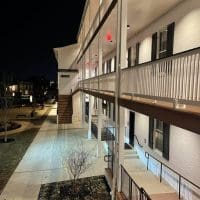 A two-story white brick building at night features a lit walkway, black shutters on windows, and white railings. External stairs lead to the upper floor. Leafless trees and landscaped areas border the walkway at Visitation Hotel Frederick MD.