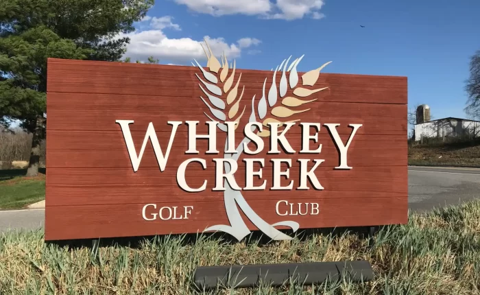 A large wooden sign reading Whiskey Creek Golf Club in white letters, with two stylized wheat stalks crossing behind the text, stands on grass beside a road in New Market MD. Trees, a blue sky with clouds, and distant farm buildings are in the background.
